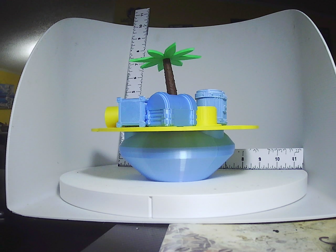 3D printed treasure island coin bank with blue ocean base, yellow disc, miniature treasure chests, and green palm tree