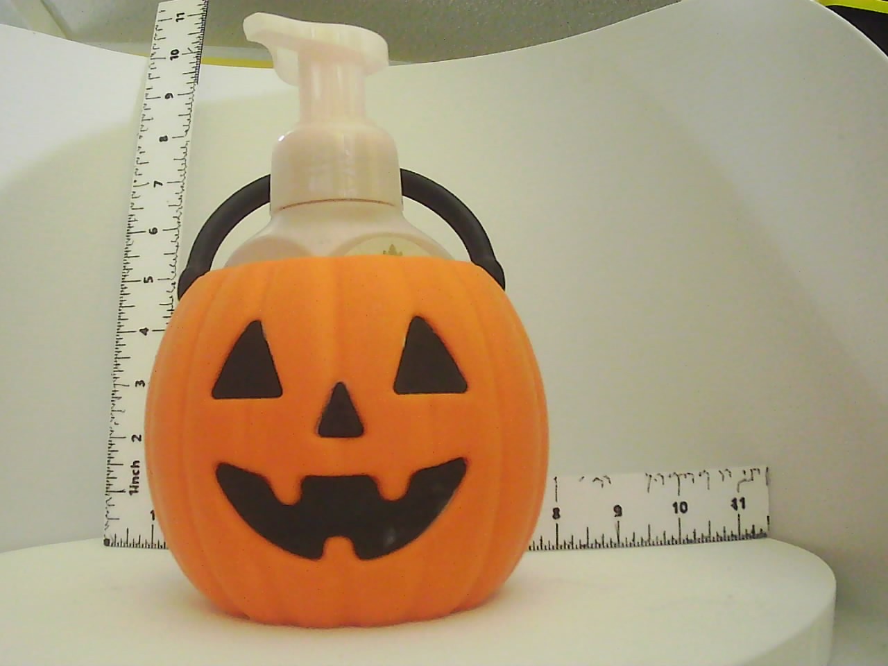 Orange jack-o-lantern soap dispenser holder with black handle cradling a pump bottle, shown with rulers for scale