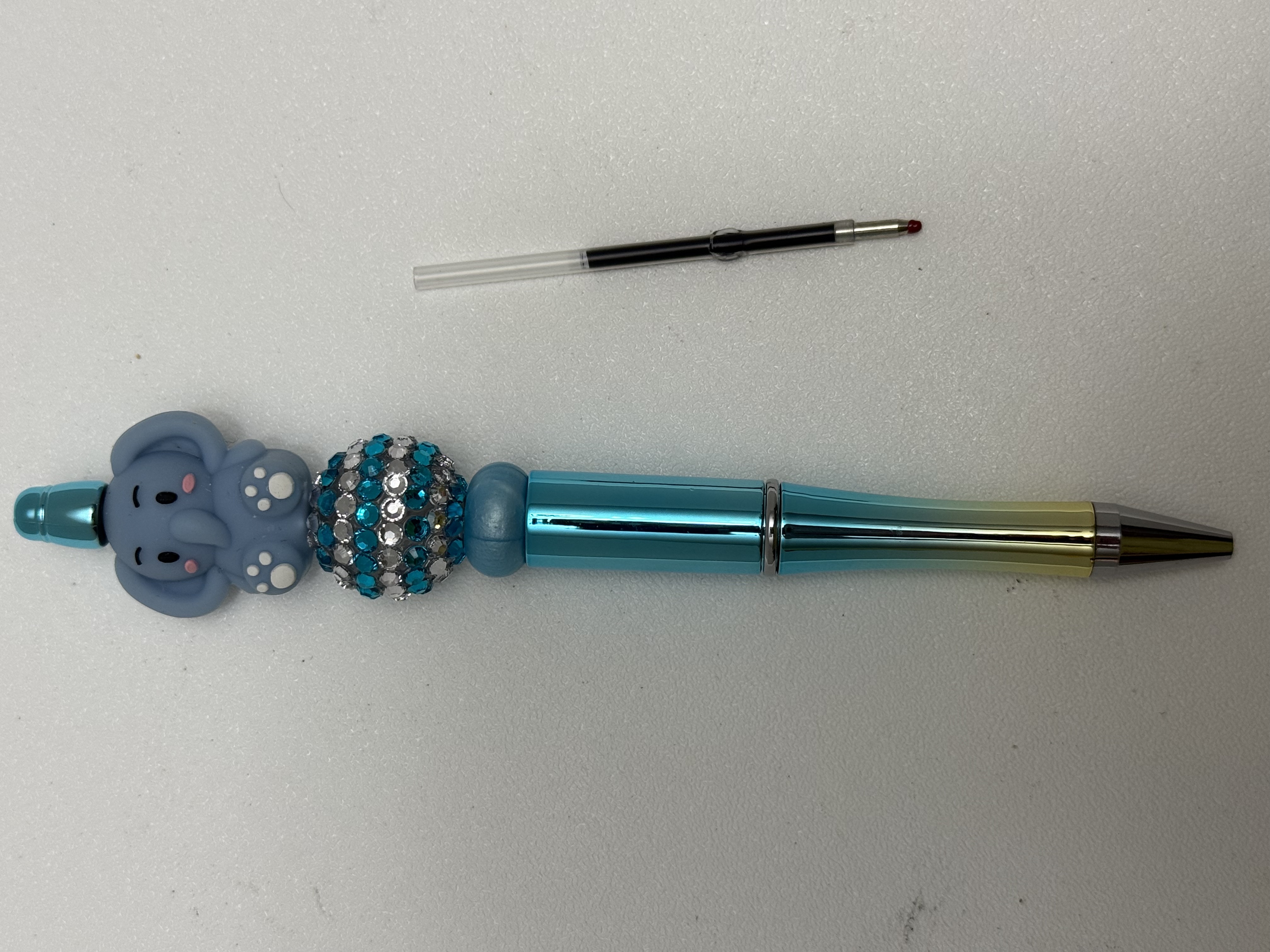 Blue gradient ballpoint pen with kawaii elephant charm, teal rhinestone bead, and spare ink refill beside it