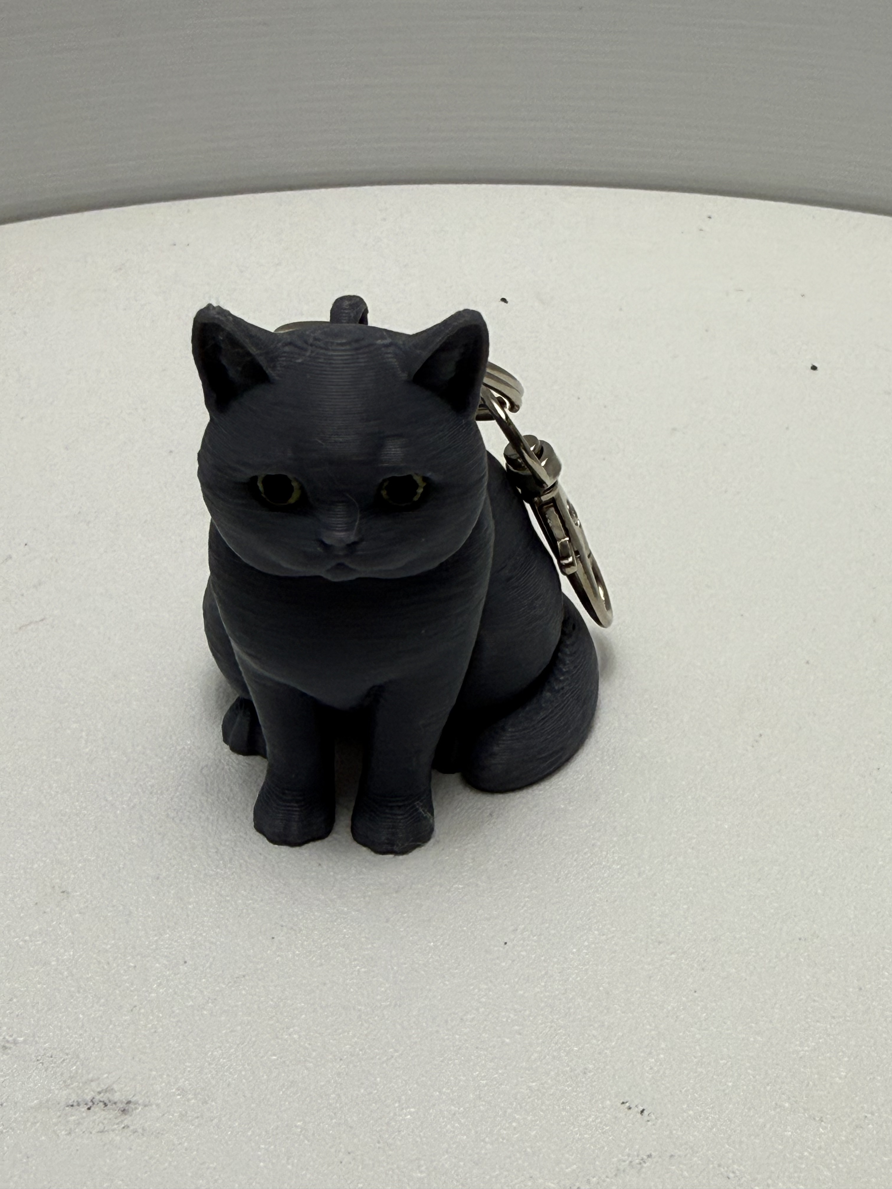 Matte black 3D printed chubby sitting cat keychain with silver lobster clasp on white surface