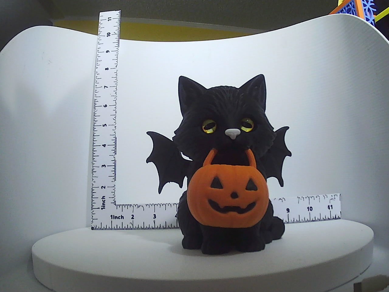 Black cat figurine with bat wings holding an orange jack-o-lantern, shown with ruler for scale