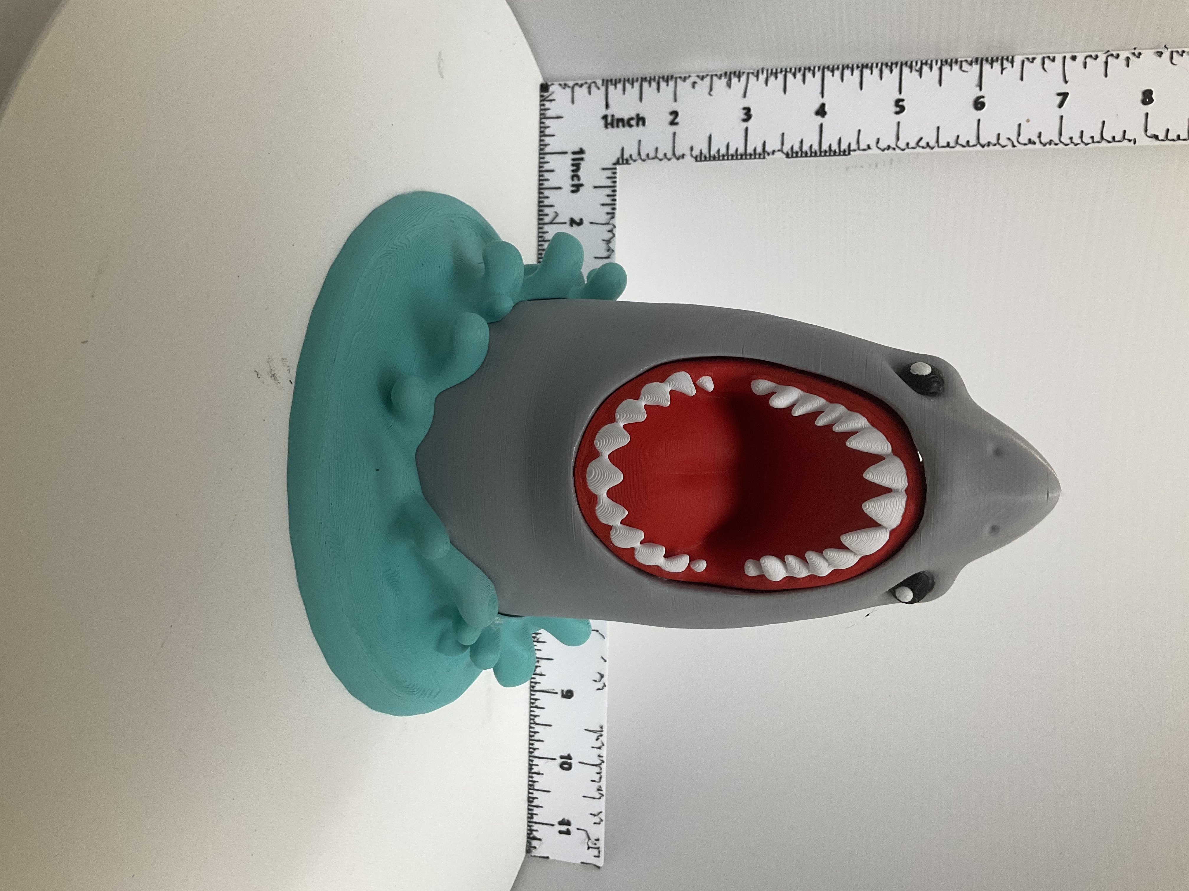 Front view of grey cartoon shark soap dispenser holder with open red mouth, white teeth, and teal wave base