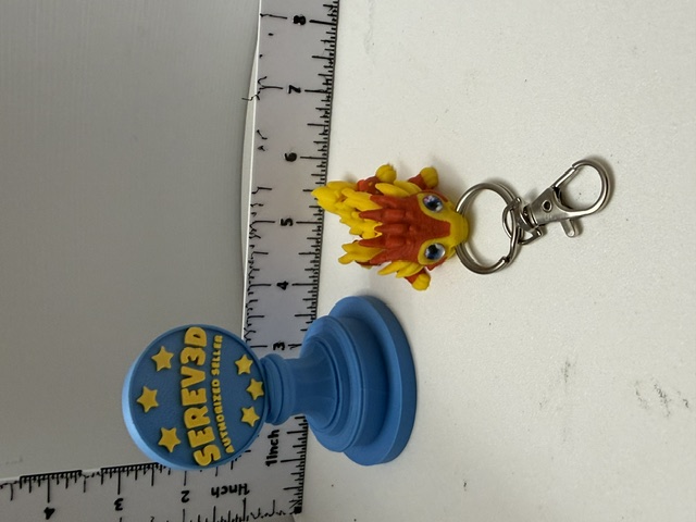 Small yellow and orange spiky dragon keychain charm with silver lobster-claw clip shown next to a ruler for scale