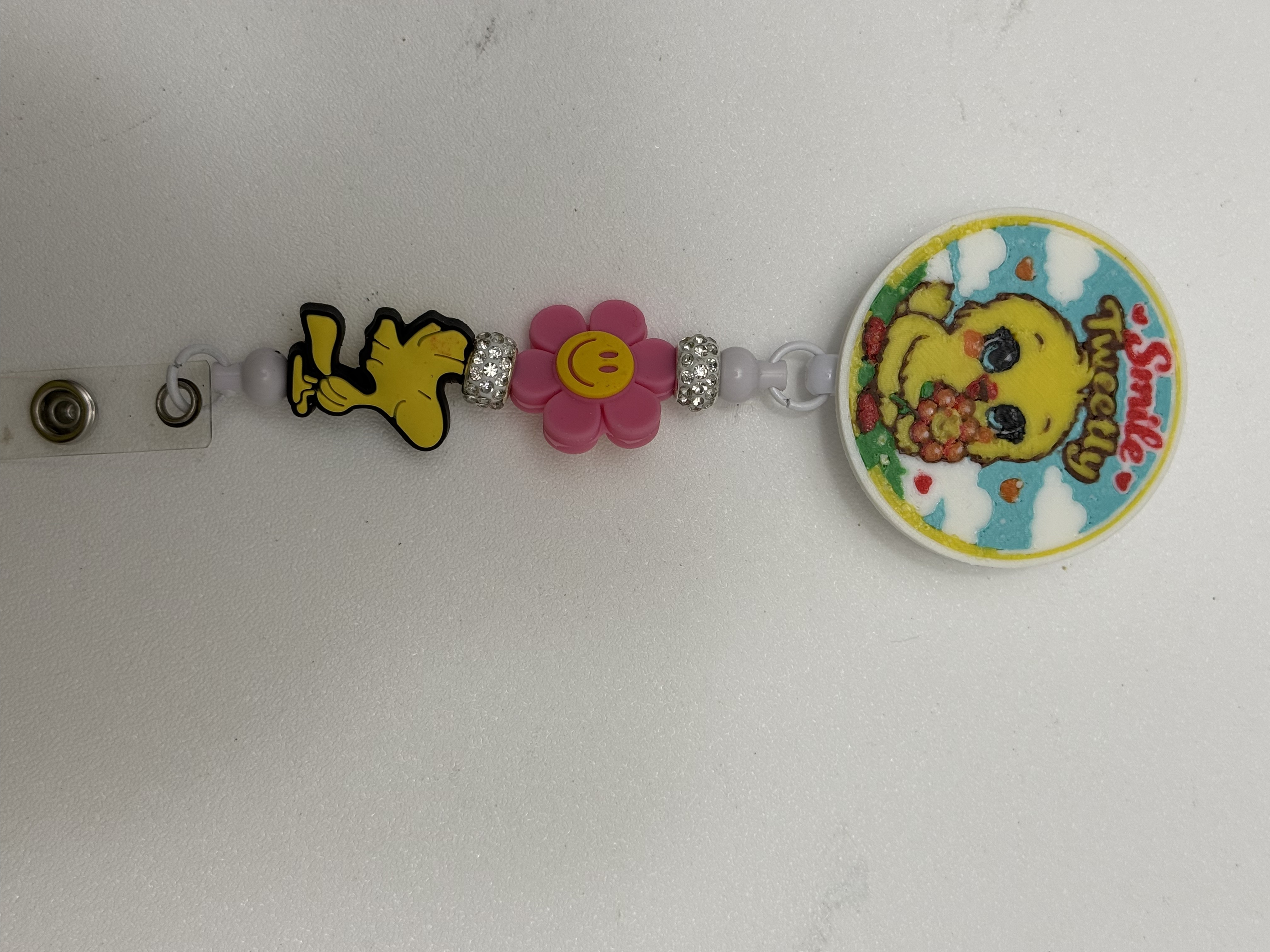 Retractable badge reel with yellow bird medallion, pink daisy bead, and rhinestone spacer beads on white background