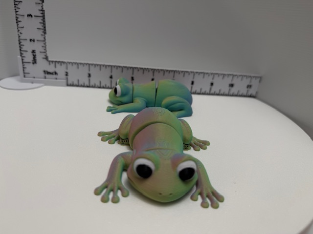 Two articulated fidget frogs with wide eyes, one matte green and one iridescent rose-teal, shown beside a ruler on a white surface