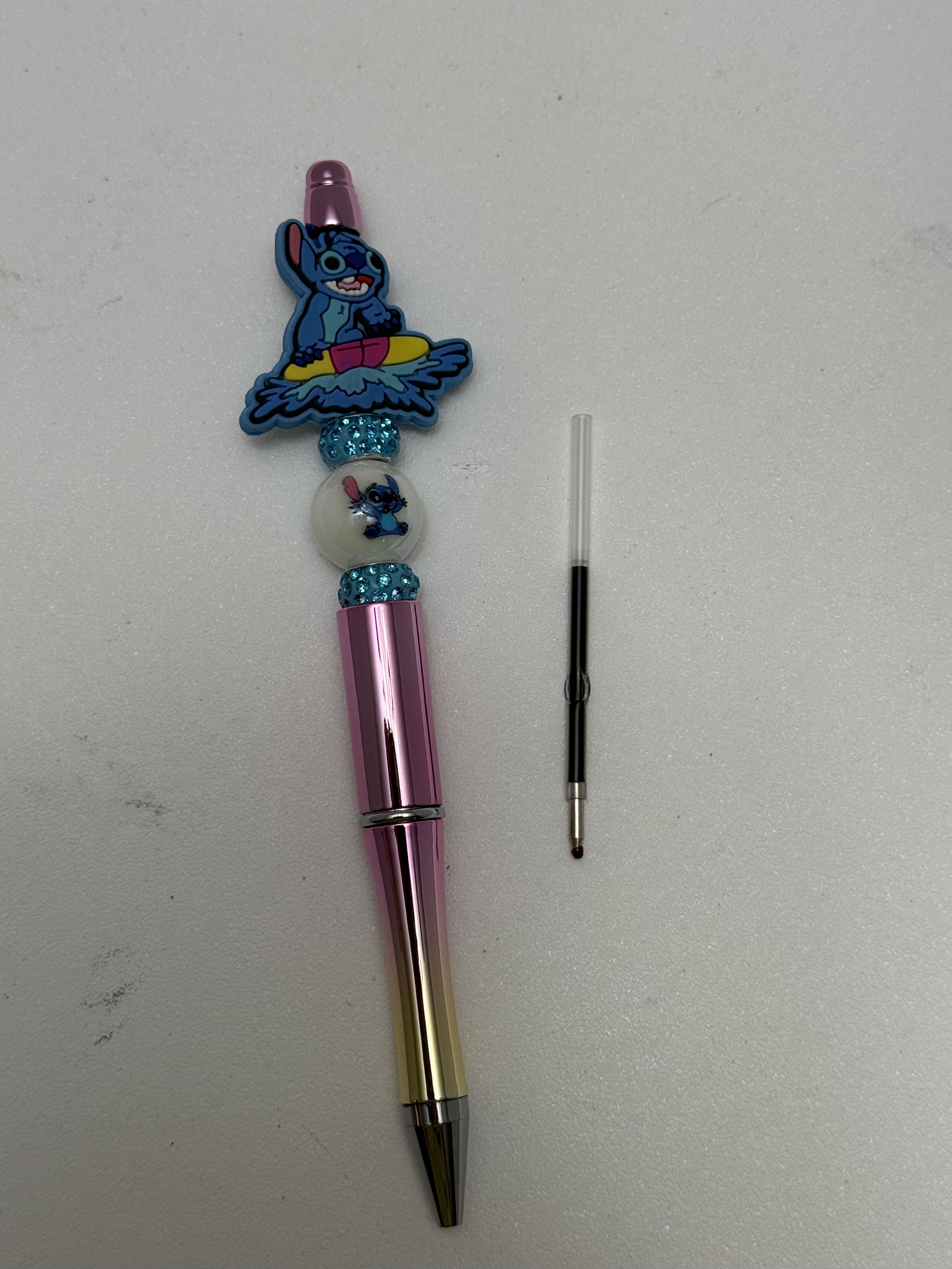 Pink metallic beaded pen with blue surfing alien charm, aqua rhinestone spacers, and a loose ink refill beside it
