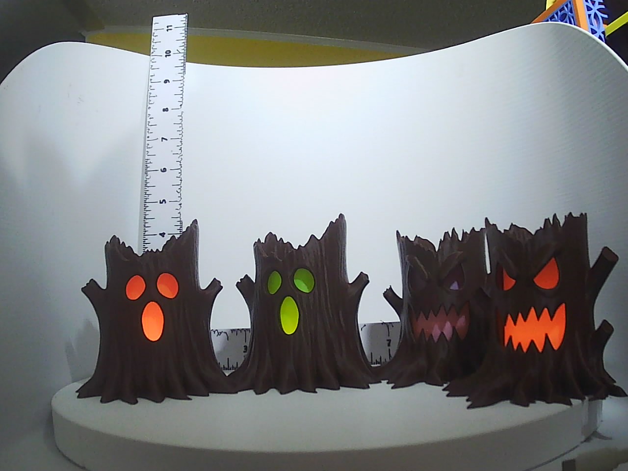 Four dark bark-textured tree stump lanterns glowing orange, green, pink, and red through spooky face cutouts