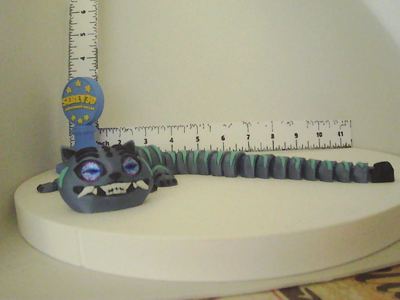 Articulated blue-grey and teal cat figure with glowing violet eyes and white fangs shown next to a ruler
