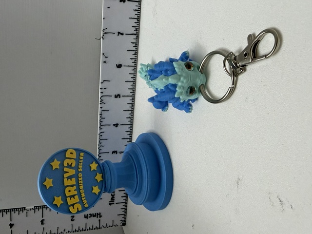 Small blue 3D-printed dragon keychain with spikes and silver lobster clasp beside a ruler
