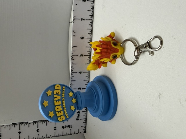 Small yellow and red dragon keychain with silver lobster clasp next to a ruler for scale