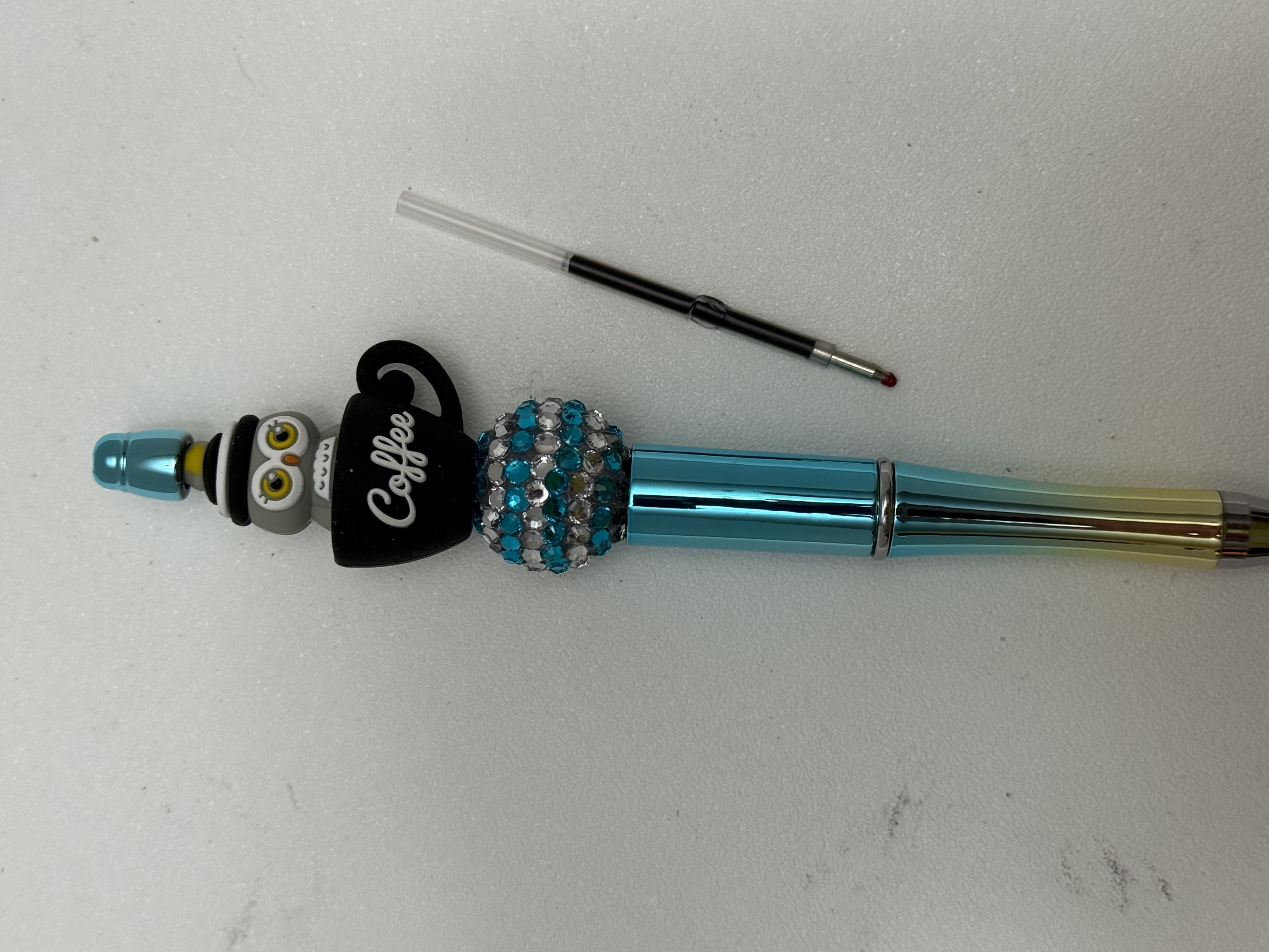 Teal beaded pen with owl and coffee cup charm beside an ink refill cartridge