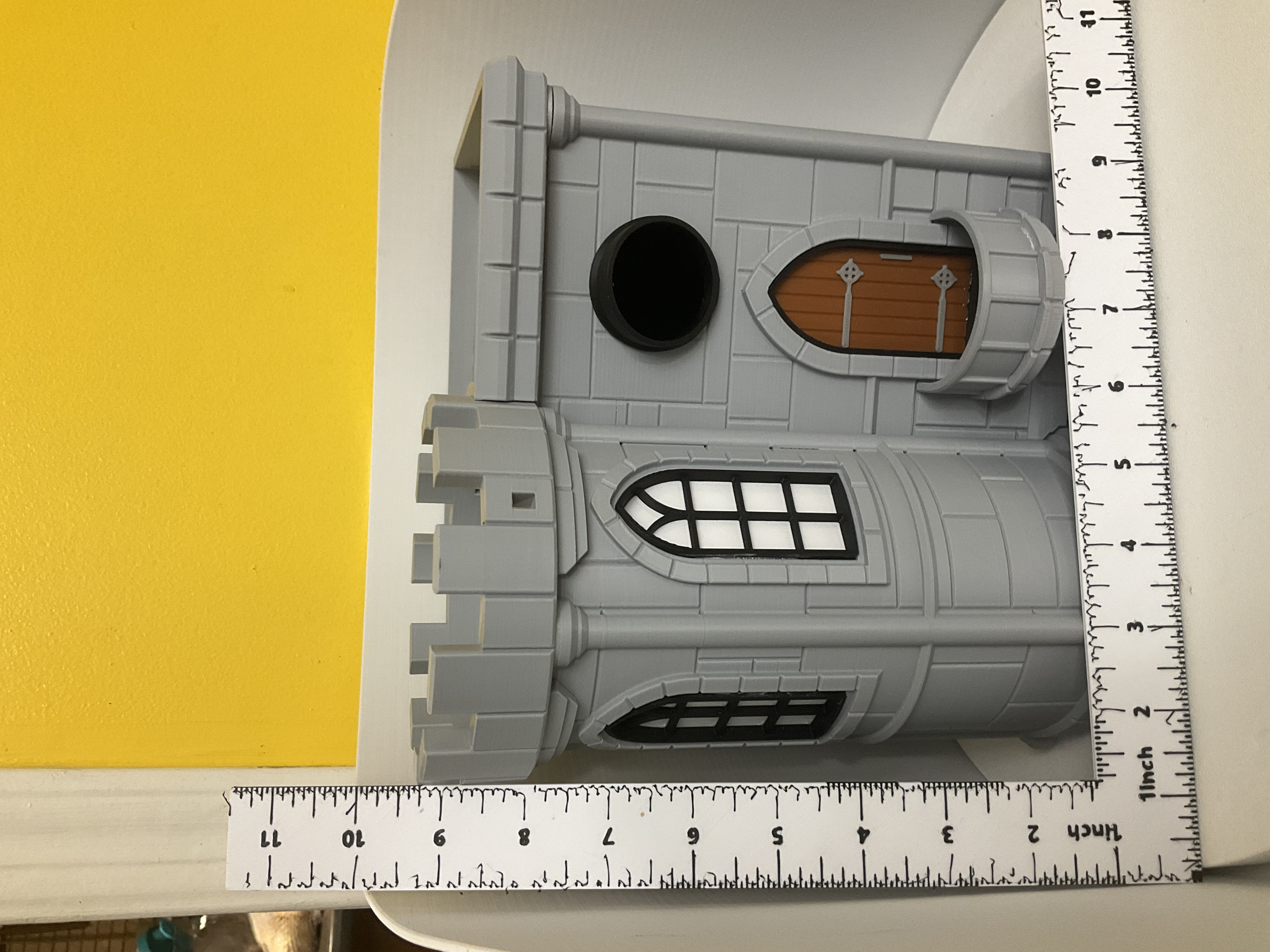 Gray castle birdhouse with round turret, arched windows, and brown wooden door shown with ruler for scale
