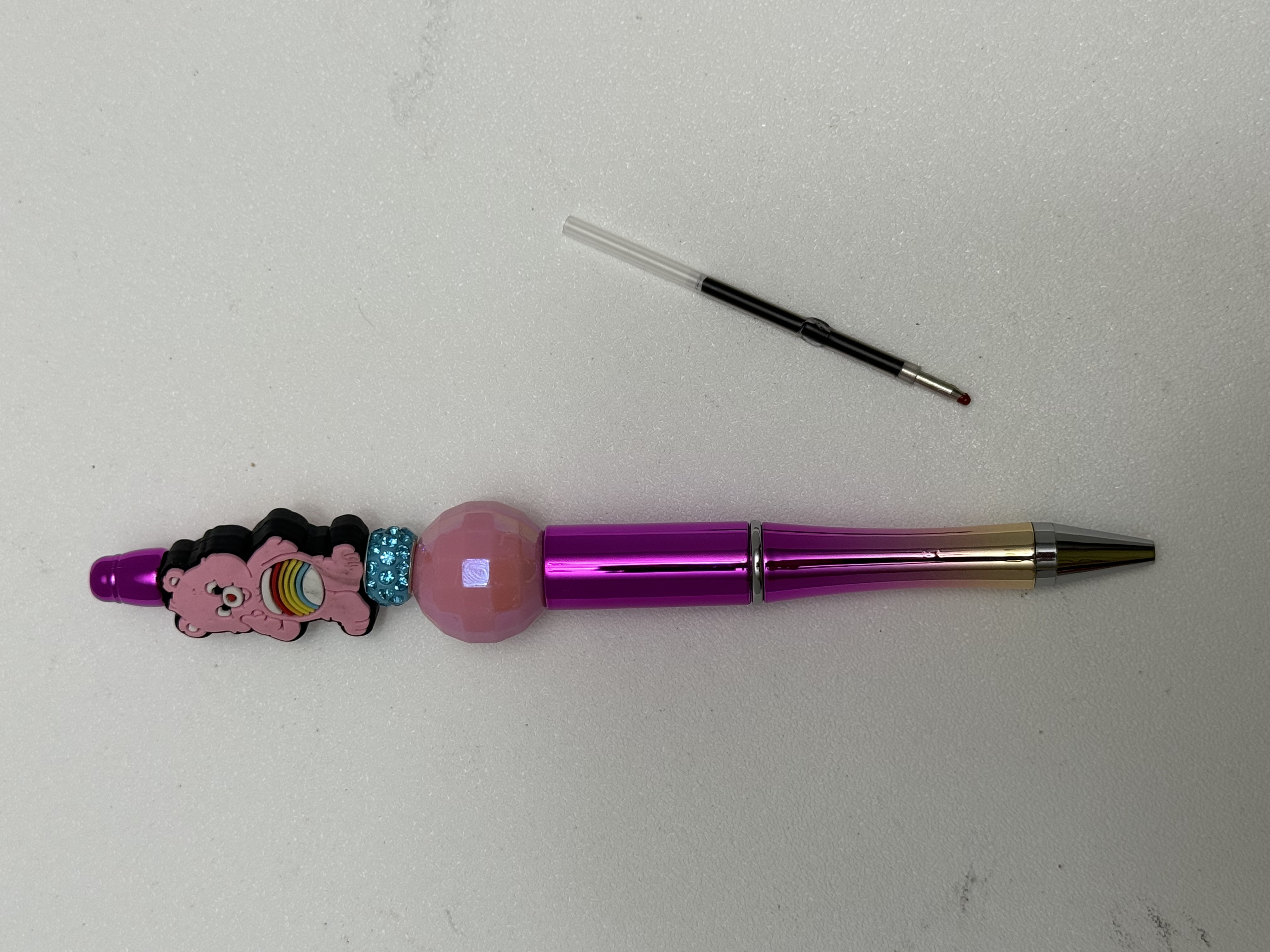 Magenta beaded ballpoint pen with bear charm, pink bead, and aqua rhinestones beside a refill