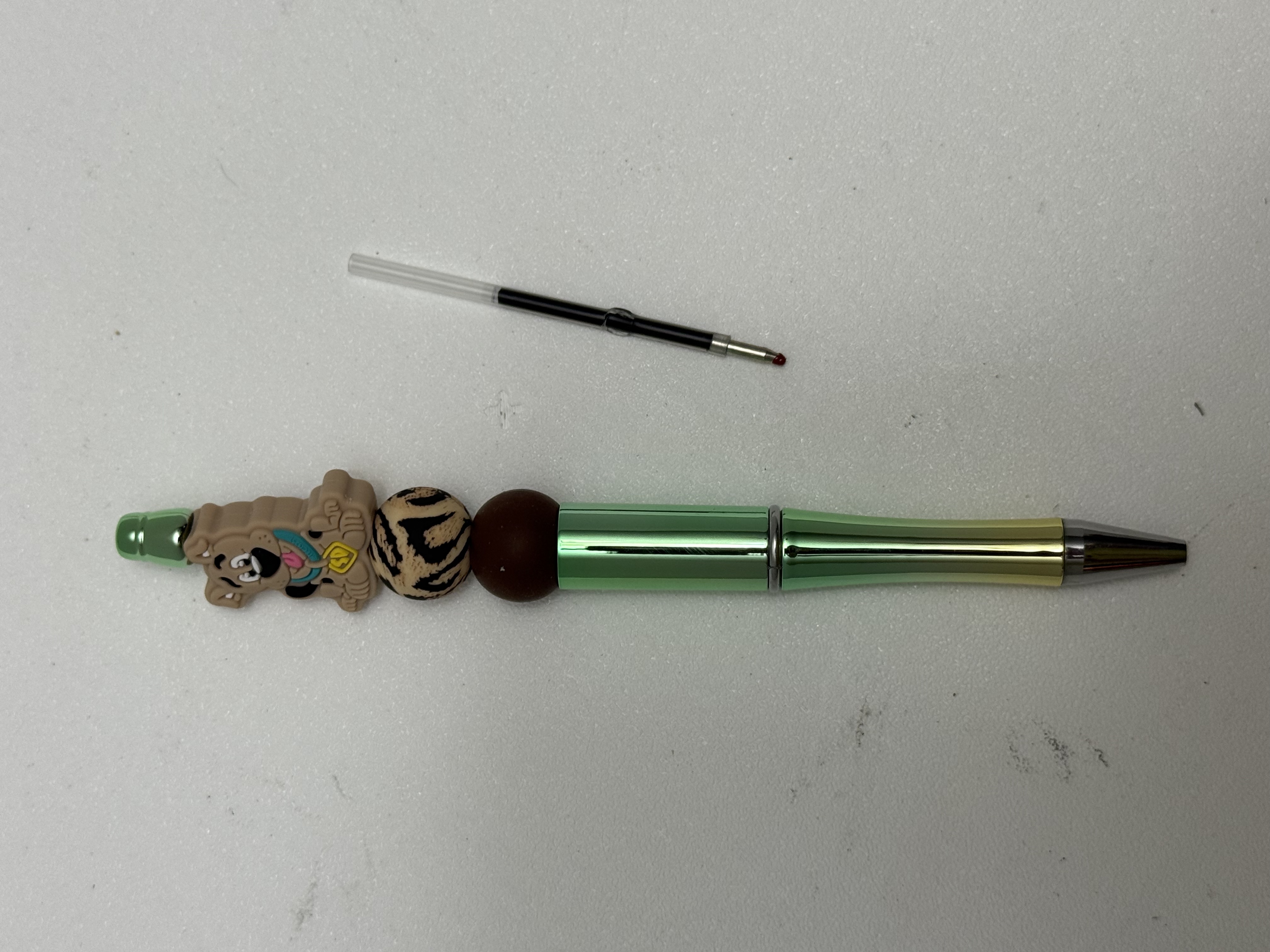 Green metallic ballpoint pen with cartoon dog charm, zebra-print bead, and dark brown bead, shown with ink refill