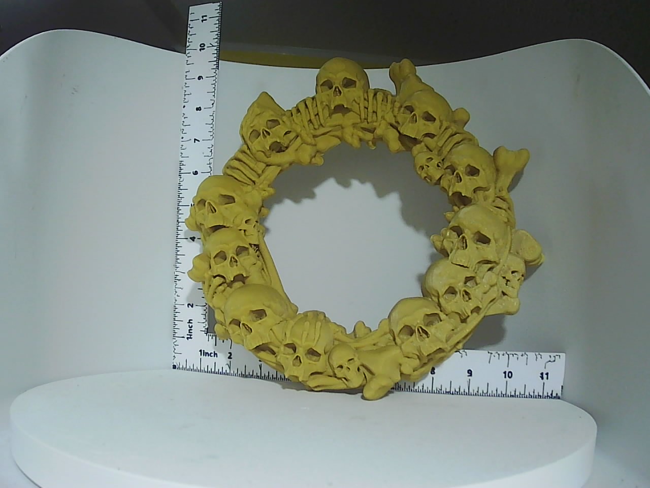 Circular wreath densely covered in printed skulls and bones in a warm bone-yellow tone, shown next to rulers for scale