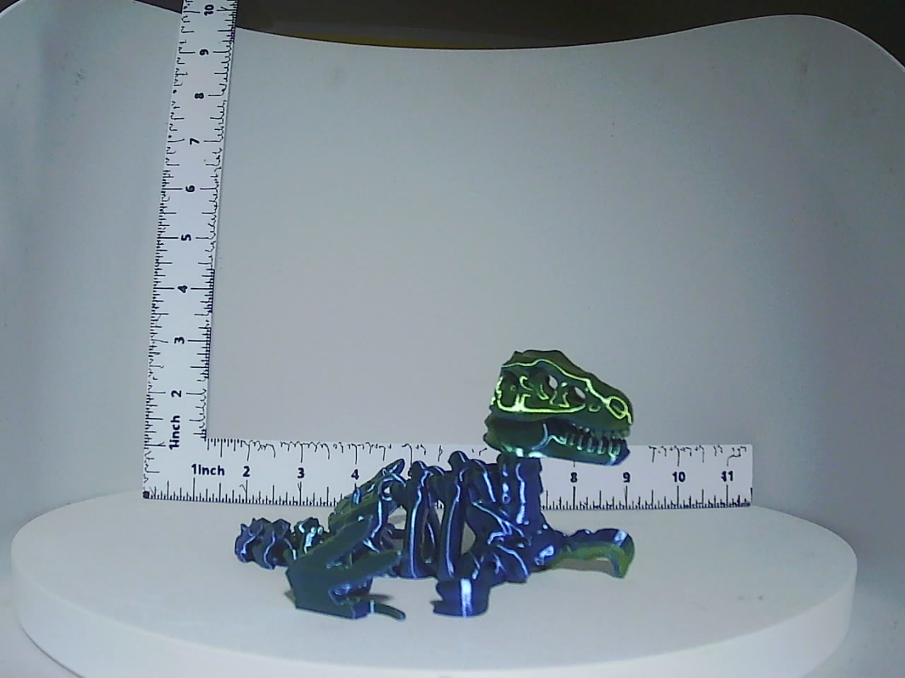 Articulated skeleton dinosaur figurine with dark body and bright yellow-green skull accents shown next to a ruler for scale