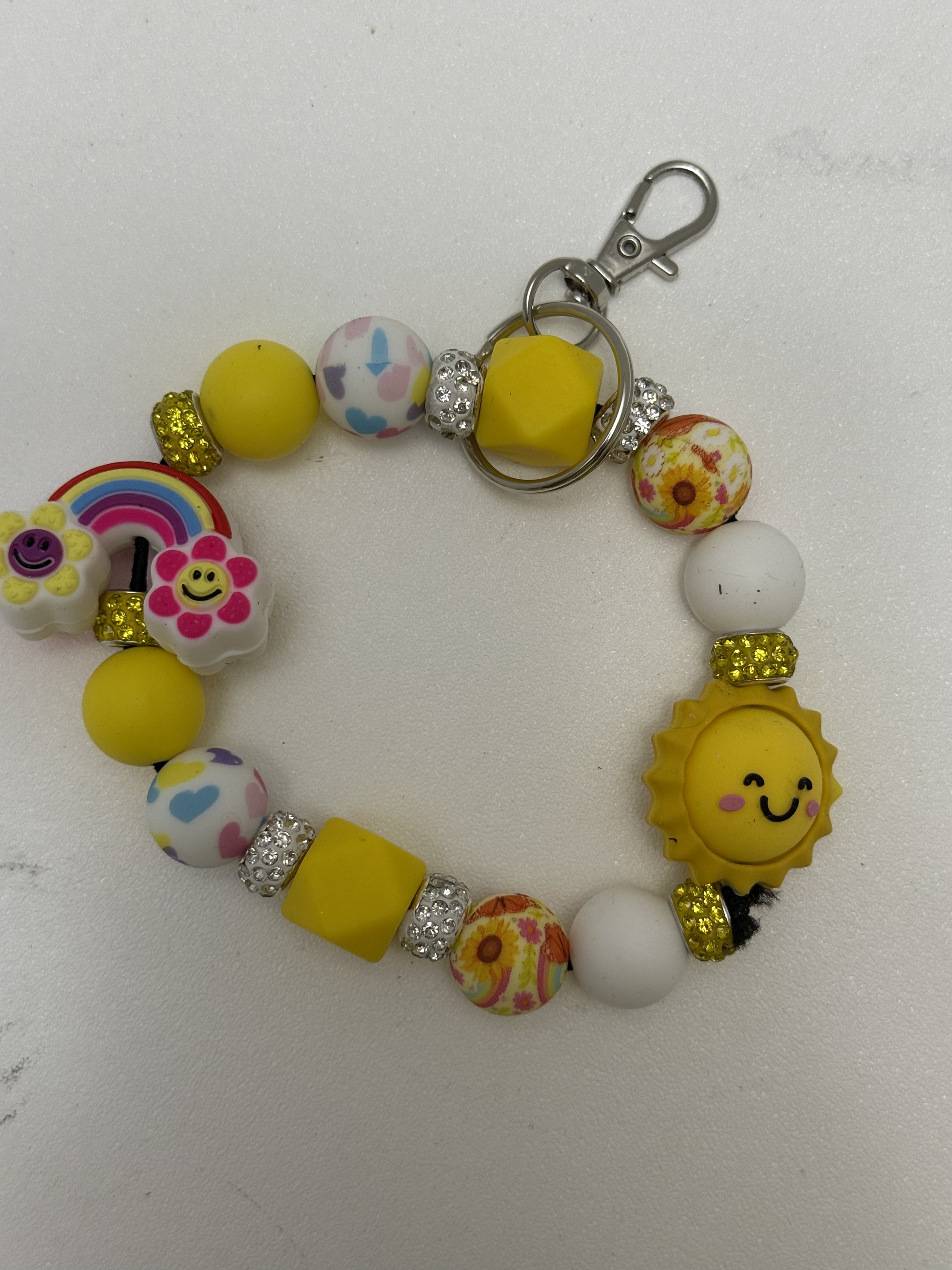 Yellow beaded wristlet keychain with smiling sun charm, rainbow focal bead, floral printed beads, and silver rhinestone rondelles on white background