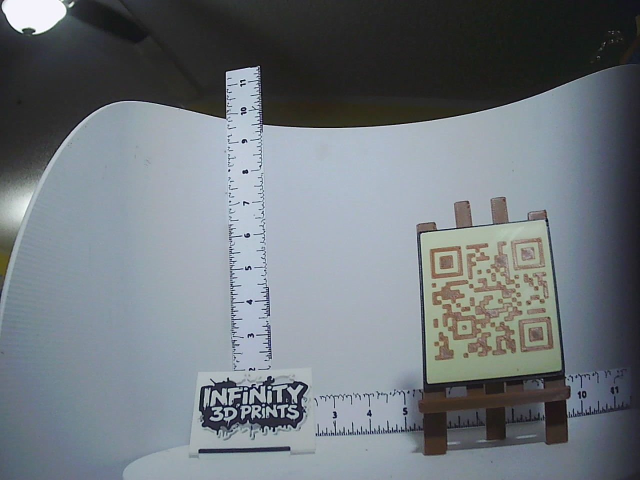 Miniature printed easel holding a cream and caramel two-tone QR code tile, shown beside rulers for scale