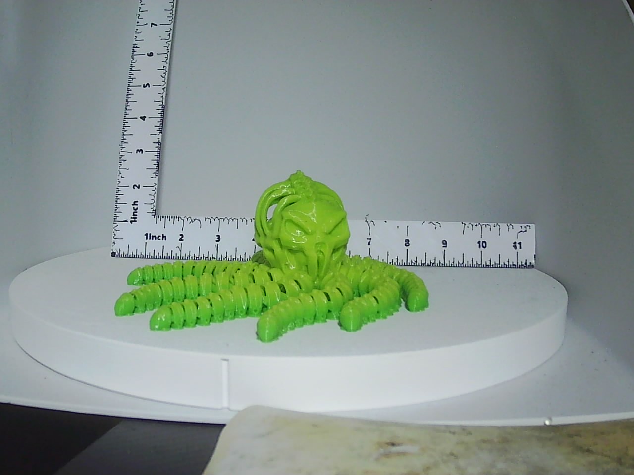 Bright green articulated skull creature with segmented tentacles displayed next to a ruler on a white turntable