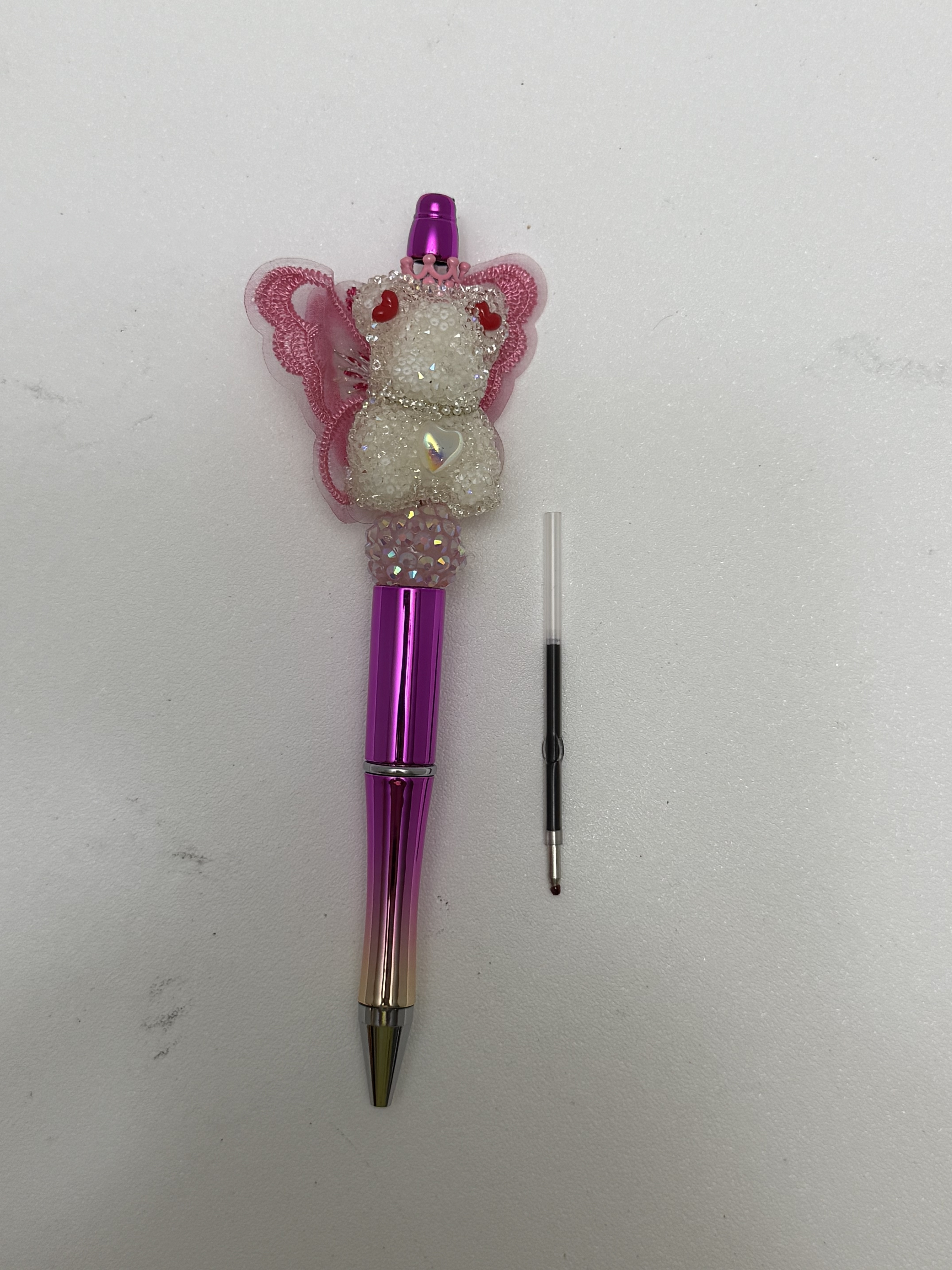 Pink ombré metallic ballpoint pen topped with a beaded bear figure wearing butterfly wings, a tiny tiara, and red heart accents, shown beside a spare ink refill cartridge