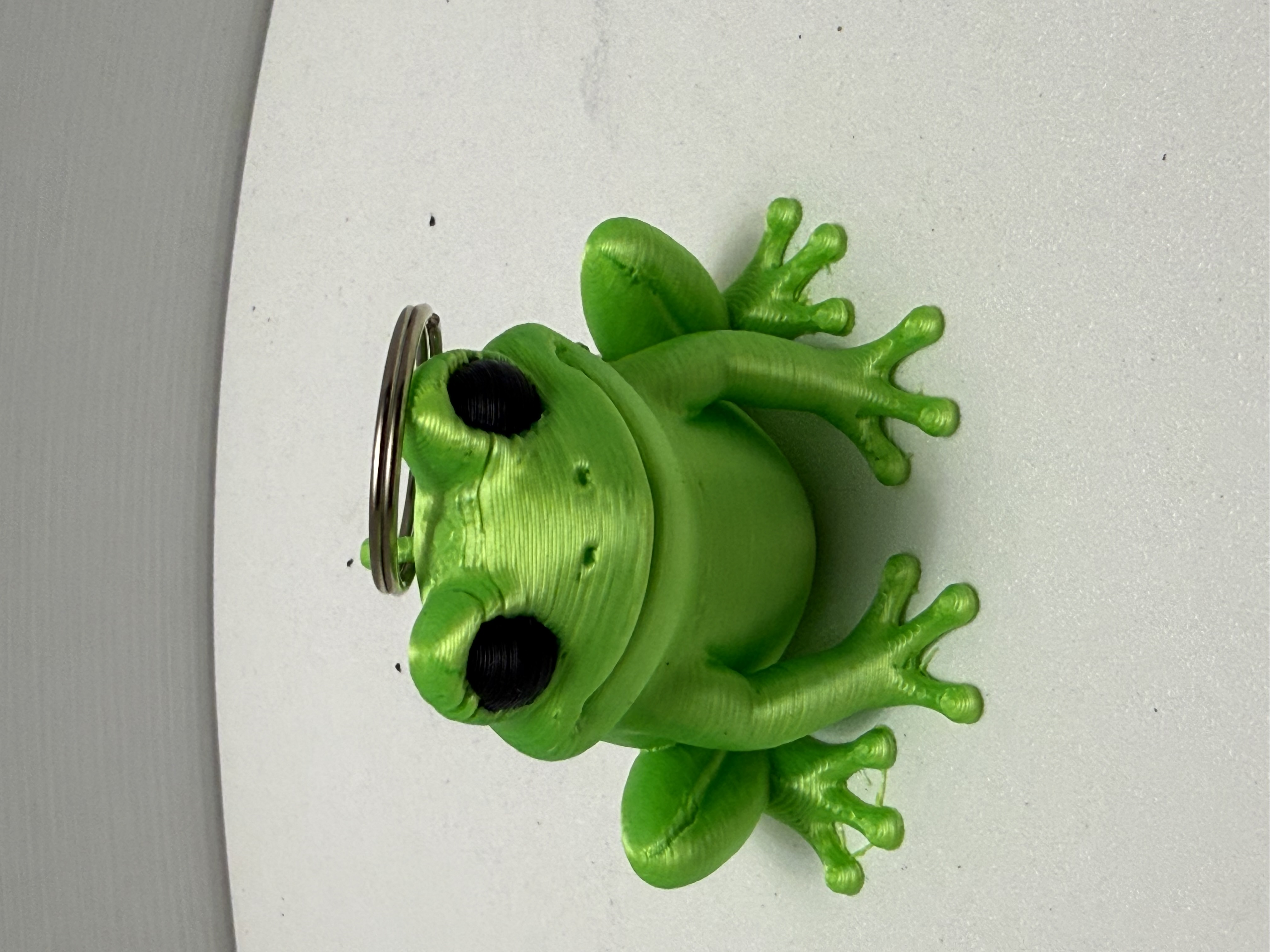 3D printed bright green frog keychain with black eyes and silver split-ring sitting on white surface