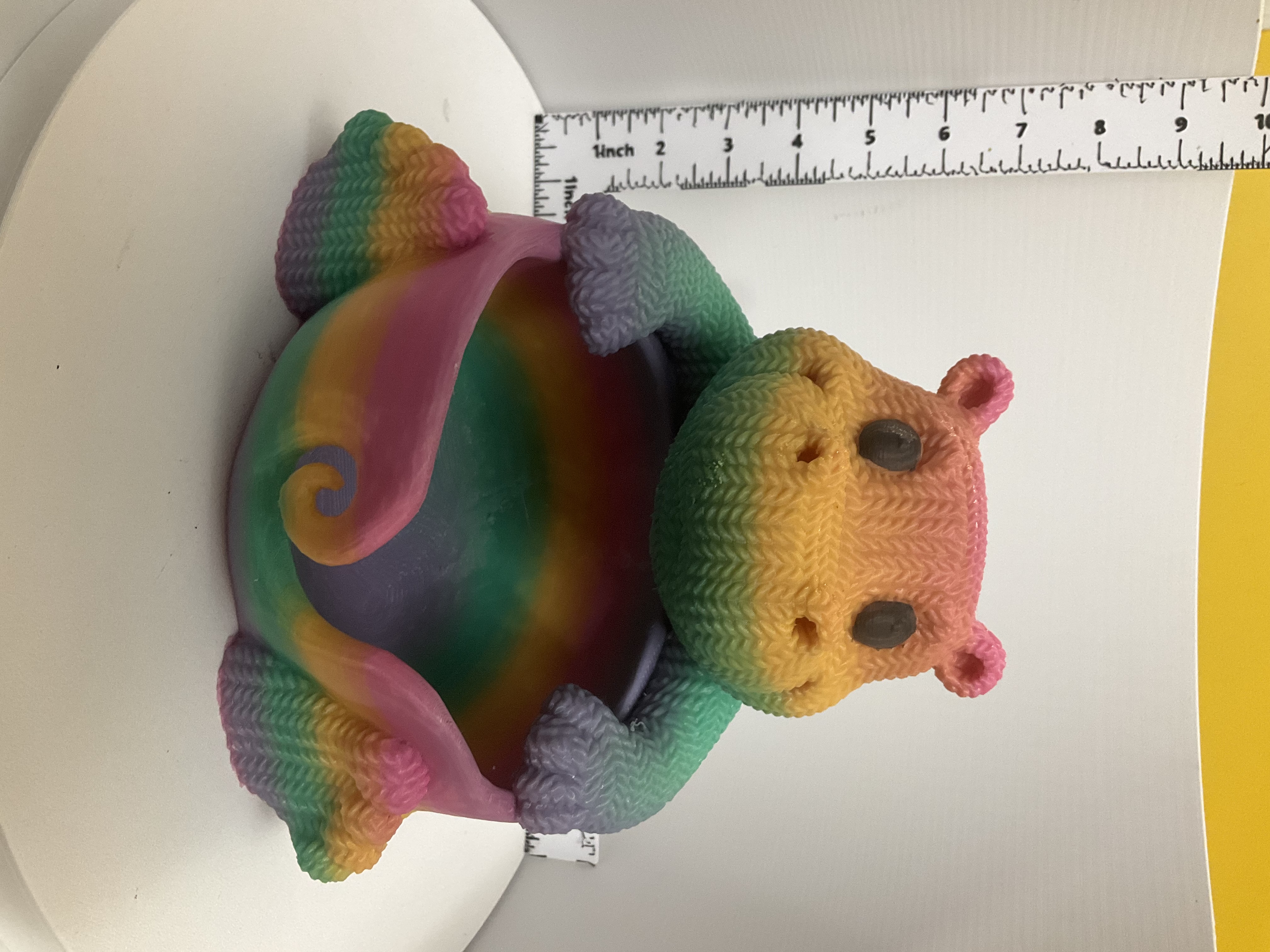 Rainbow gradient knit-texture hippo figurine cradling an open decorative bowl, shown beside a ruler for scale