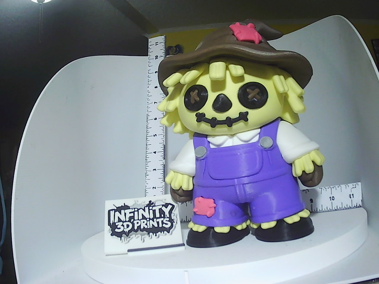 Chibi scarecrow figure in purple overalls with button cross eyes and stitched smile, next to a ruler for scale