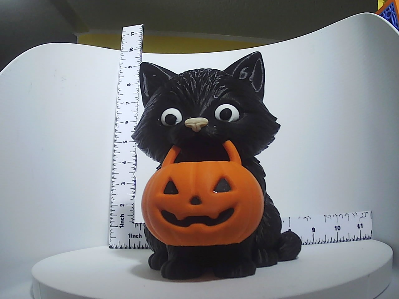 Textured black cat figurine holding an orange jack-o-lantern, shown with ruler for scale