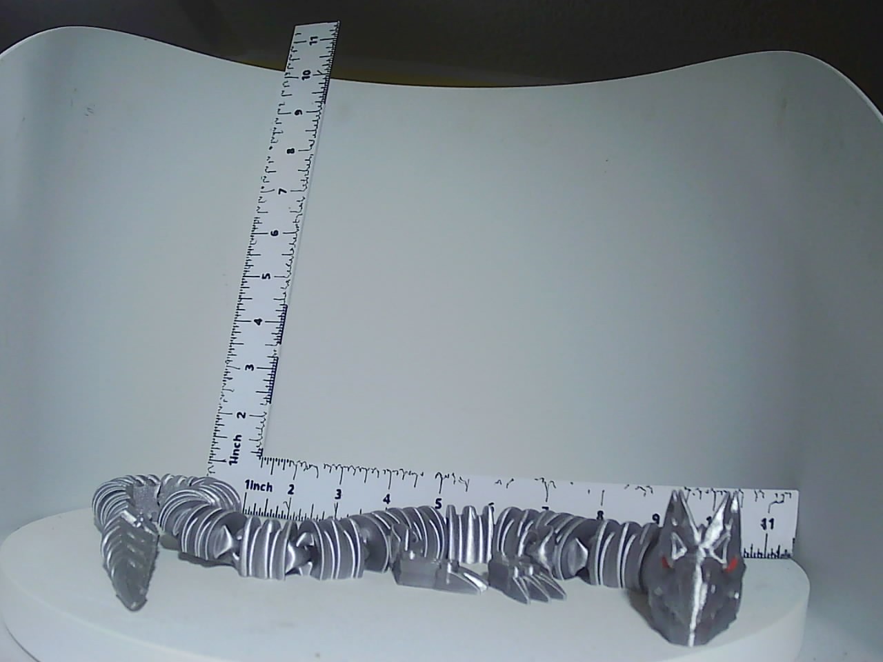 Metallic silver articulated dragon with red eye accents shown alongside a ruler for scale