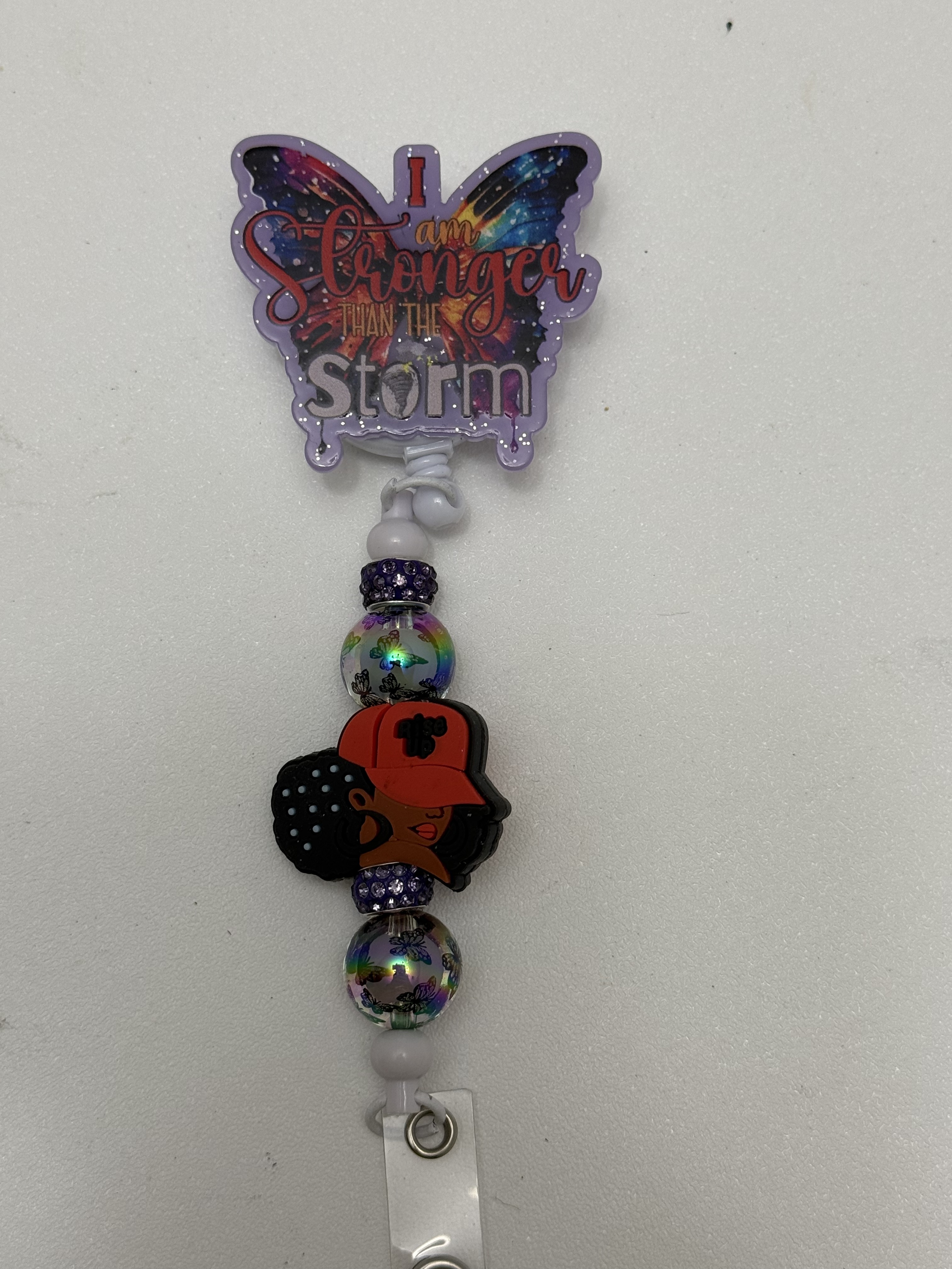 Lavender butterfly badge reel with stronger than the storm text, iridescent beads, and natural hair silhouette charm