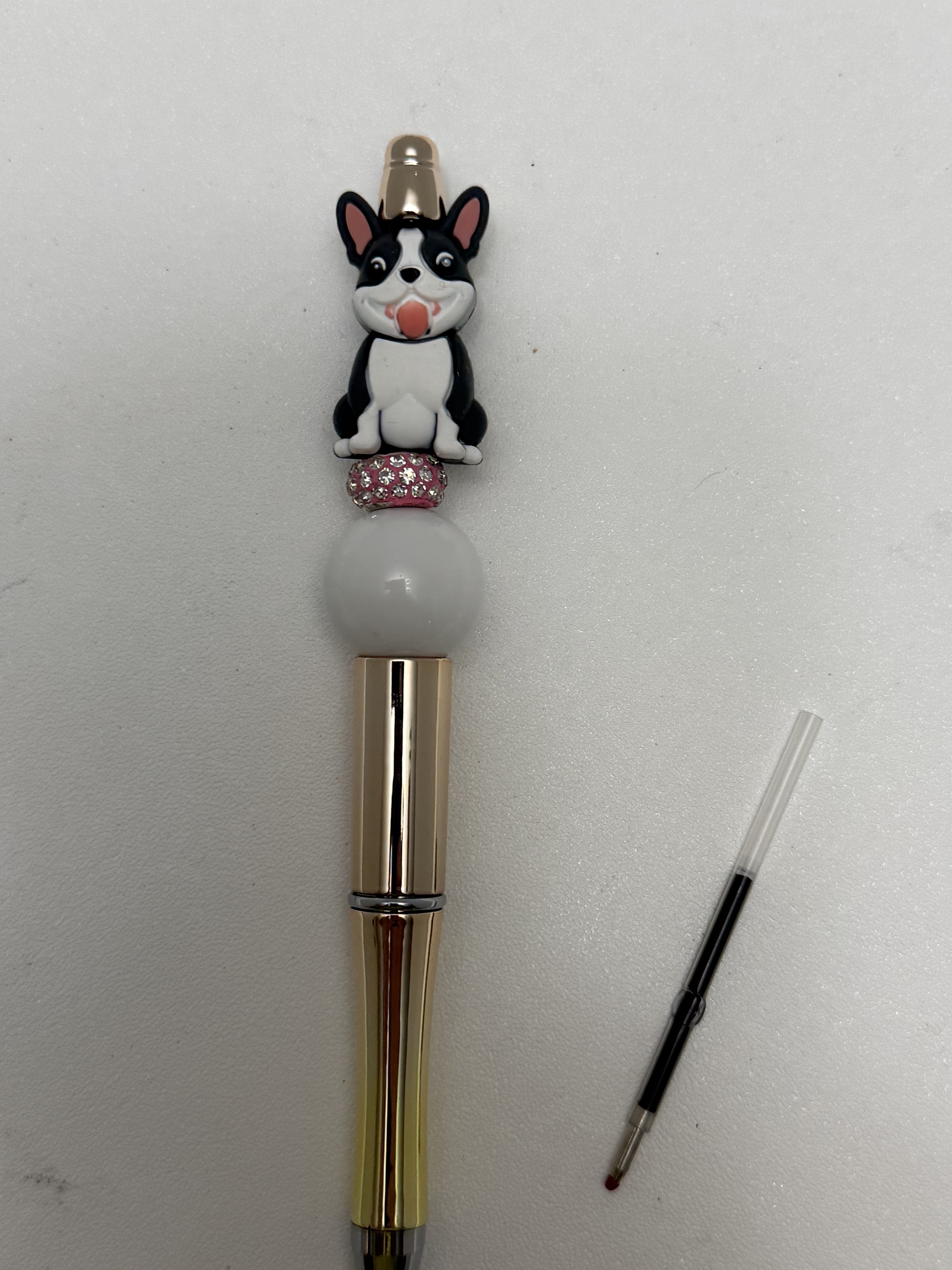 Gold-tone ballpoint pen with cartoon dog topper, pink rhinestone bead, grey round bead, and ink refill shown beside it