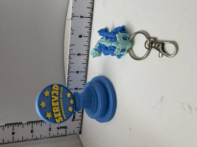 Small aqua-blue 3D-printed dragon head keychain with silver lobster clasp shown next to a ruler for scale