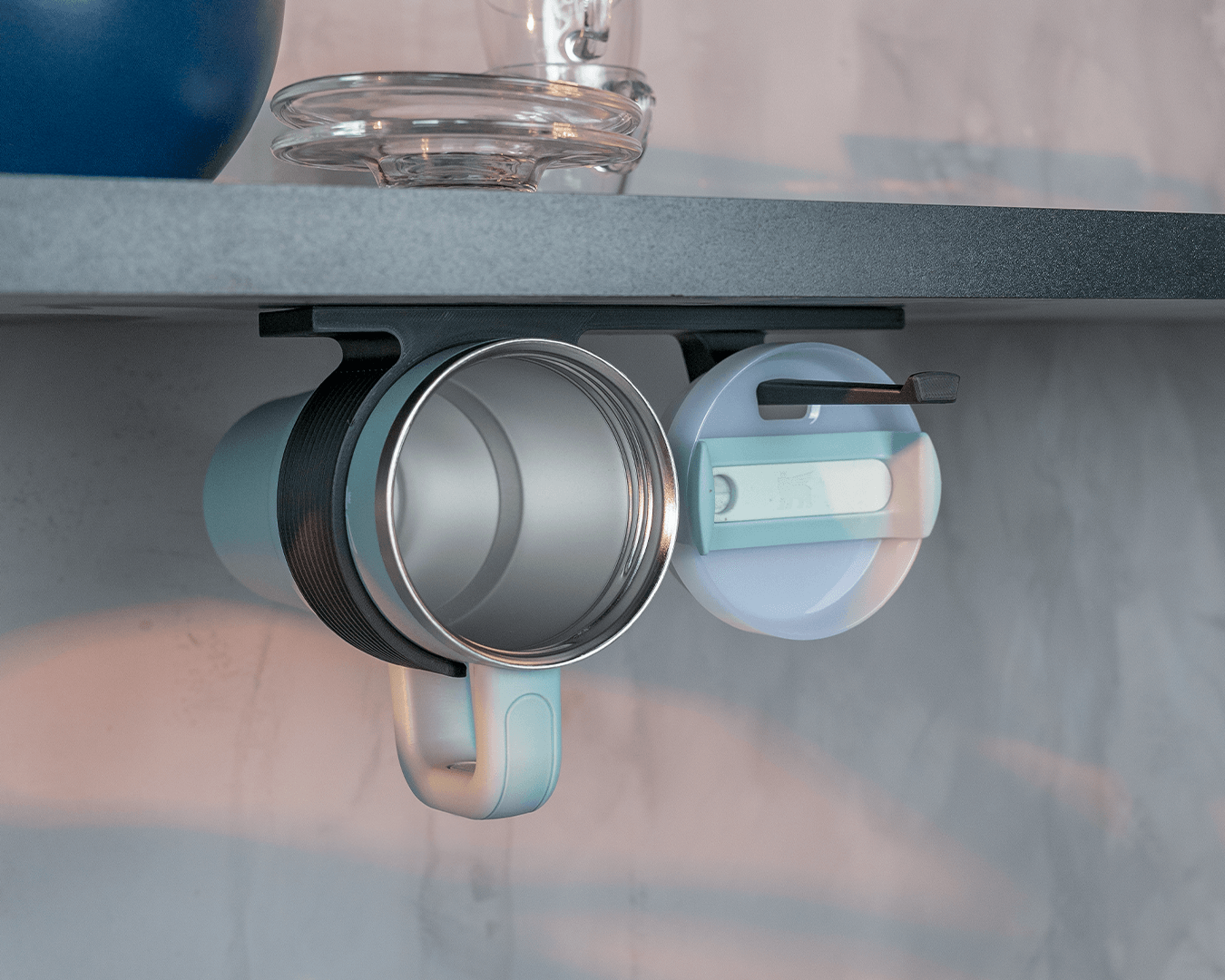 Two under-counter cup holders mounted beneath a dark shelf holding a mug and tumbler lid