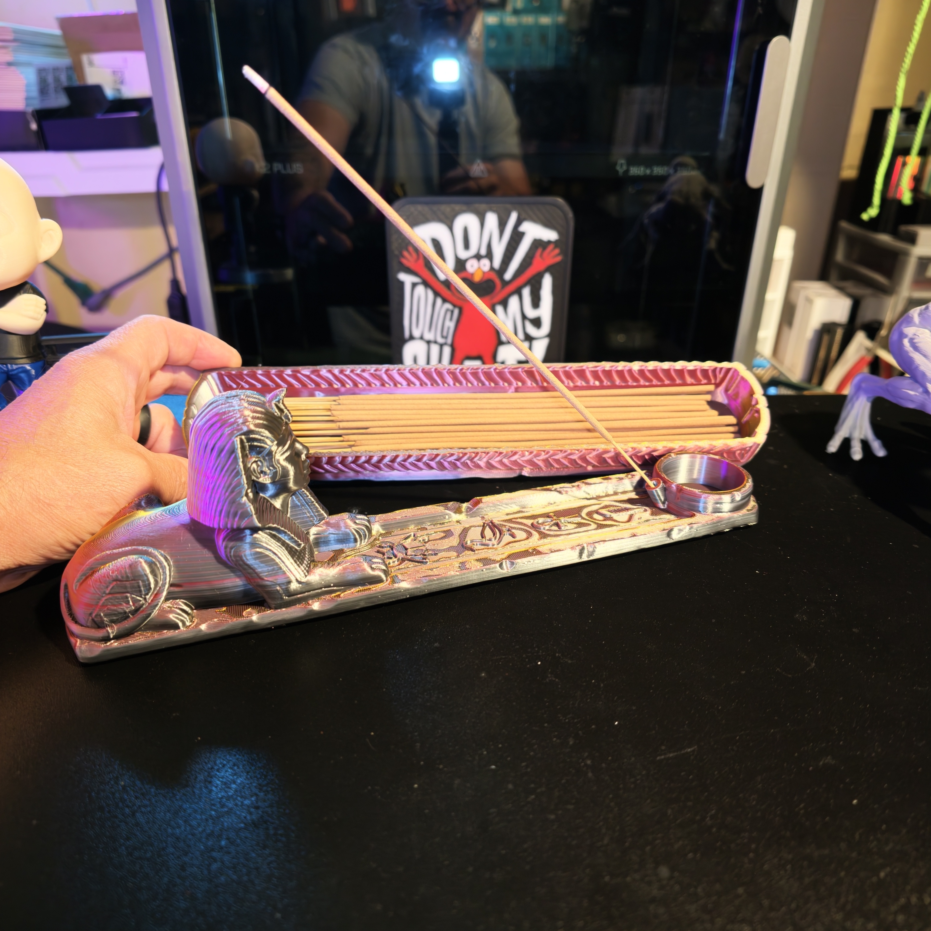 Iridescent 3D printed sphinx incense burner tray held above a black surface, loaded with incense sticks and one stick burning in the circular holder