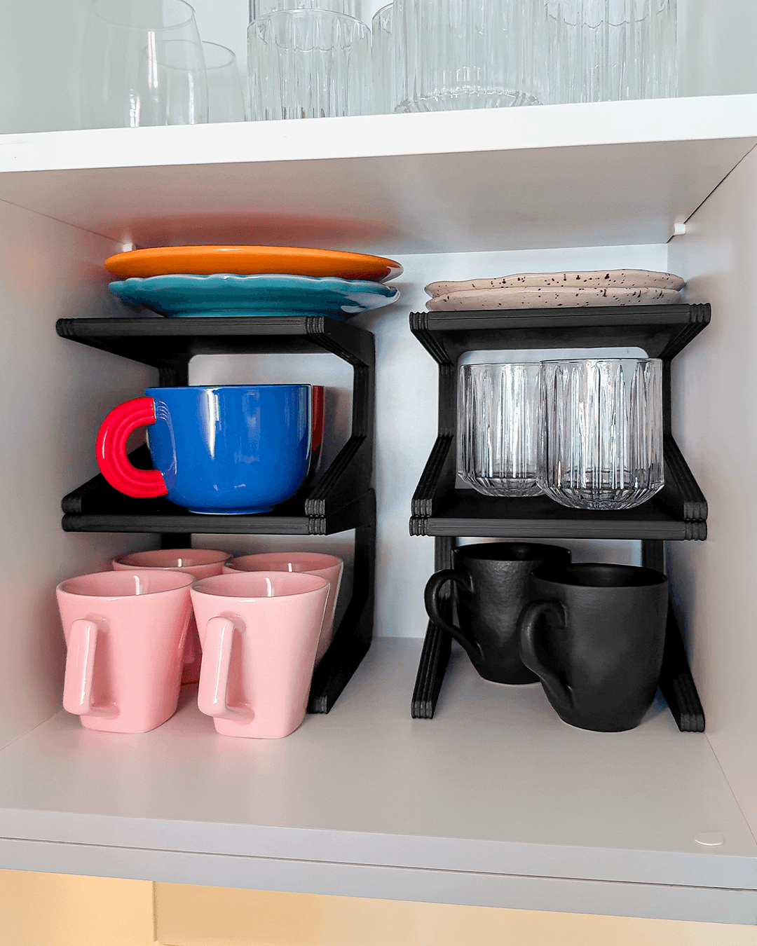 Two black 3D-printed two-tier cupboard risers holding mugs, plates, and glasses inside a white kitchen cabinet